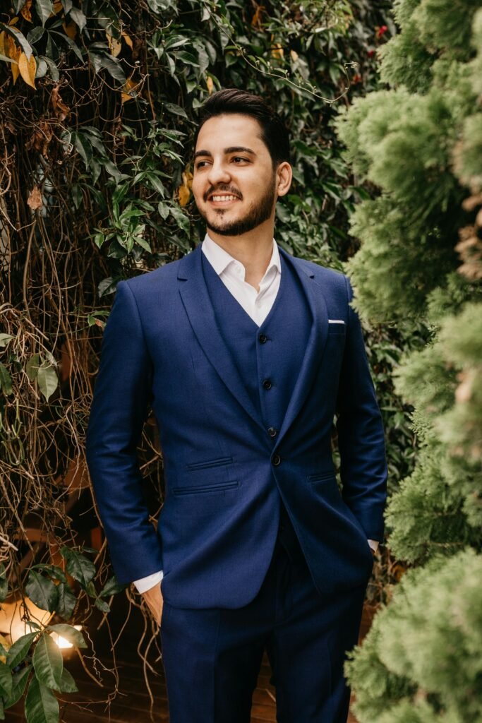 Charming groom in a stylish blue suit posing amidst greenery, exuding elegance and happiness.