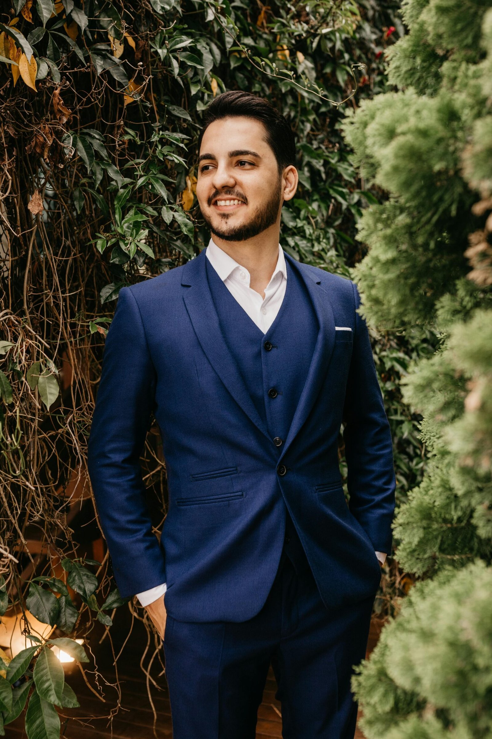 Charming groom in a stylish blue suit posing amidst greenery, exuding elegance and happiness.