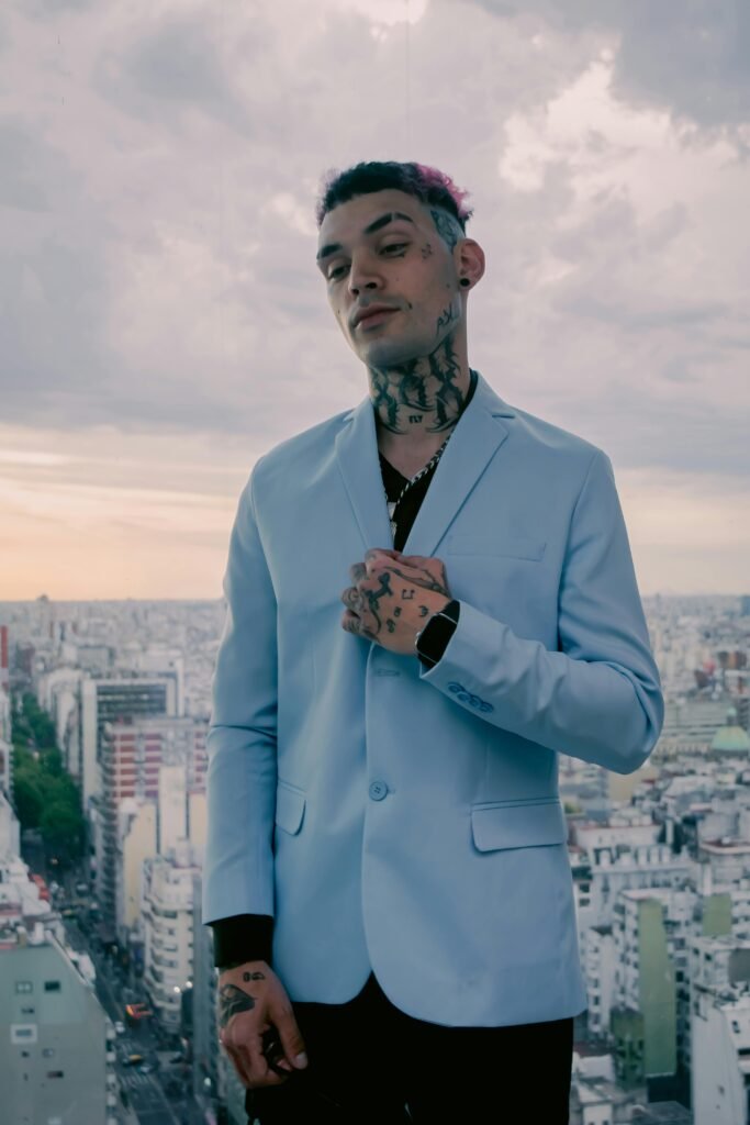 A fashionable man with tattoos poses in a light blue suit, skyline backdrop.